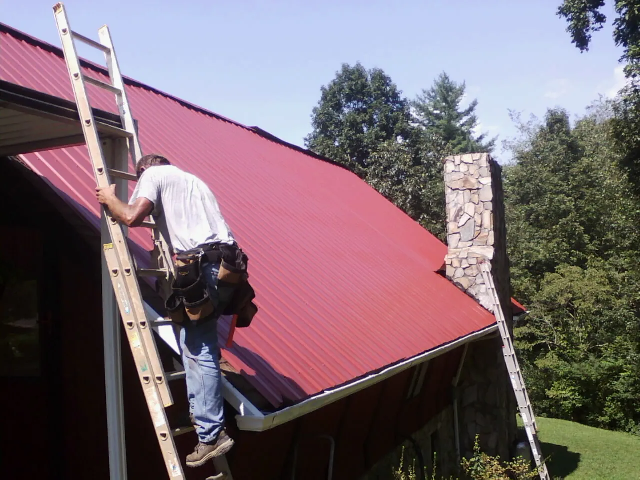 Roofer on a ladder beside a red metal roof and stone chimney — Soffit & Fascia Repair by Roofing Brothers in Sauk Rapids